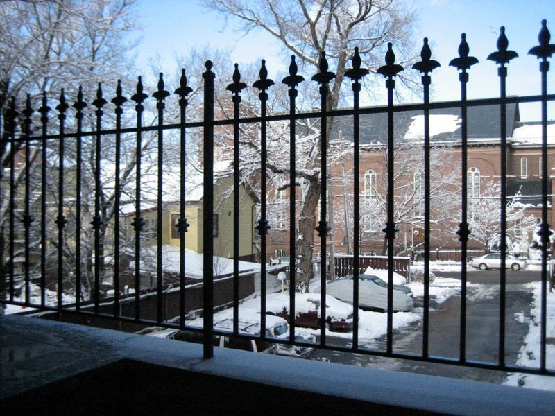 A black metal fence with pointed finials overlooks a snow-covered yard, parked cars, and buildings in winter.