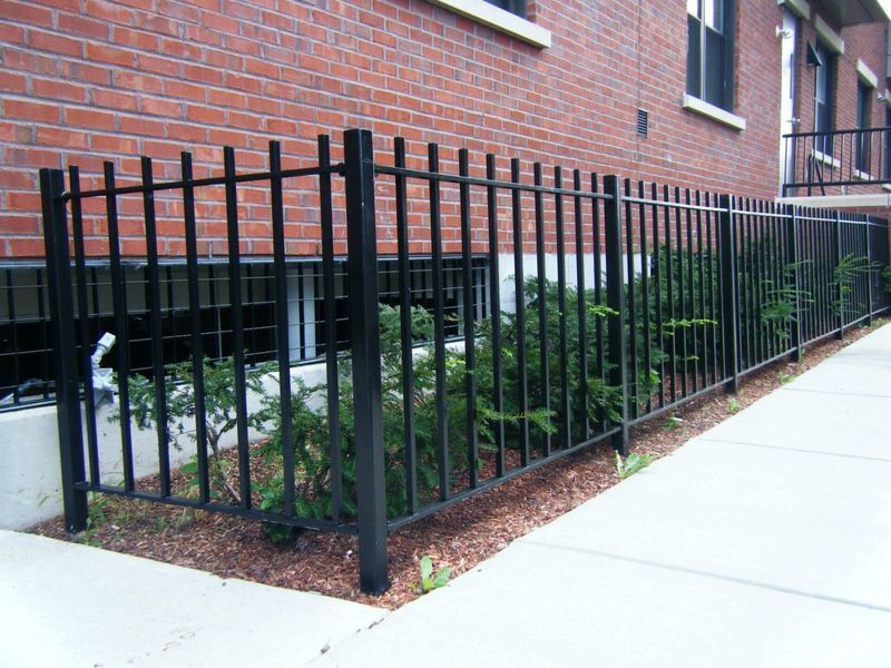 Black metal fence bordering a small garden bed against a red brick building with basement windows.