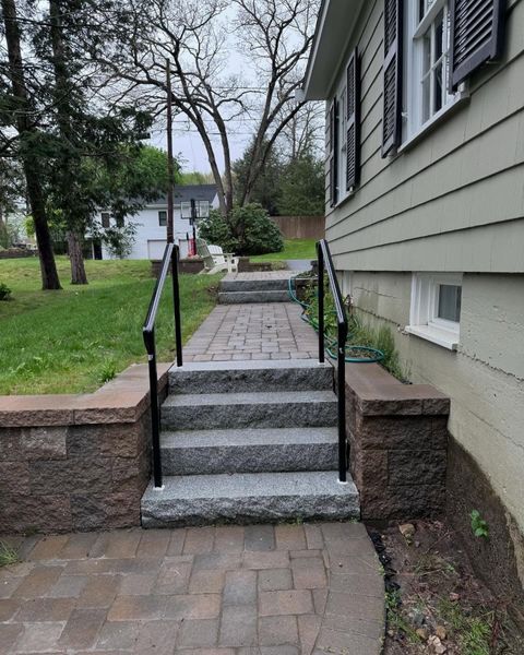 Gray stone steps with black metal handrails leading to a paved walkway beside a house exterior.