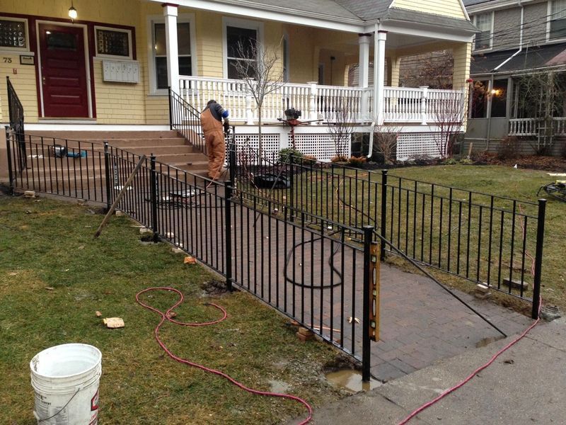 A construction worker installs black metal fencing along a paved walkway leading to the front porch of a yellow house.