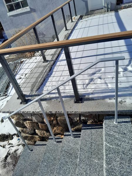 Outdoor stone steps with a gray metal handrail lead to a platform with a deck railing, partially covered in snow.