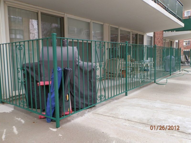 A green metal railing encloses an apartment patio containing a grill covered by a dark protective tarp.