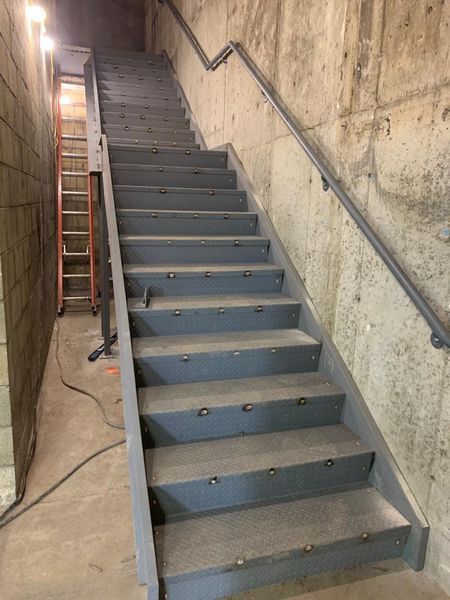 A gray metal staircase with a handrail alongside a concrete wall, with an orange ladder standing in the background.