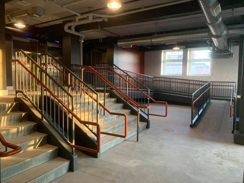 An indoor staircase with brown handrails beside a metal-railed ramp in an industrial-style building.