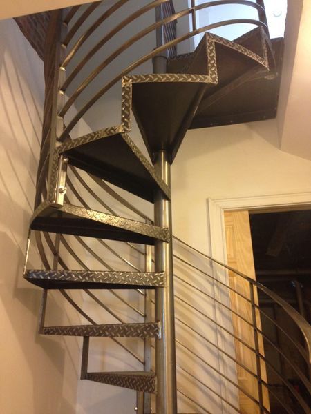 A spiral staircase with metal diamond-plate steps and a railing, installed indoors against a corner wall.