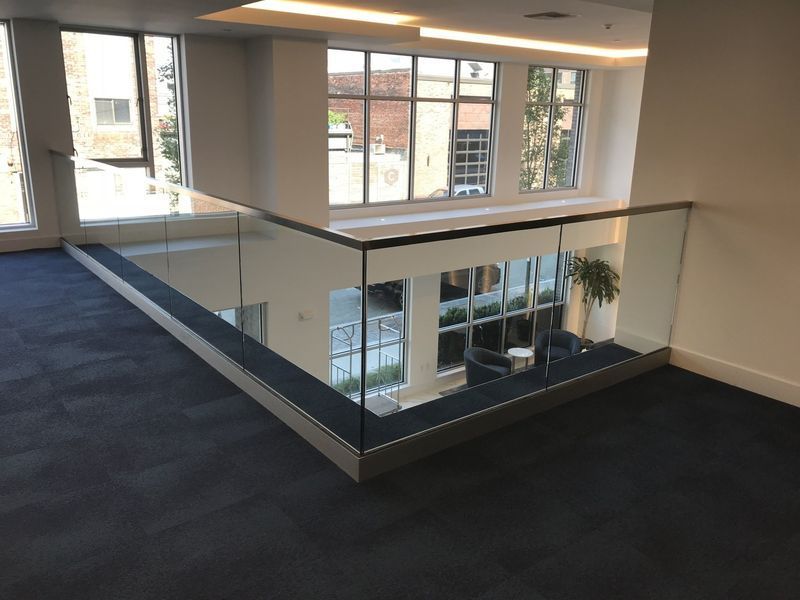 An interior view of a modern building mezzanine with a dark blue carpeted floor and a glass railing overlooking a lobby.