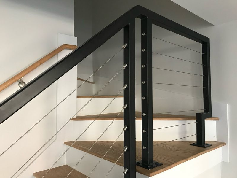 Modern staircase with black metal posts, cable railings, and wooden steps, featuring an additional wooden handrail.