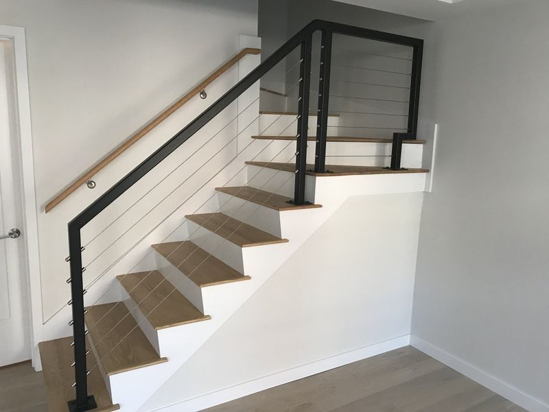 Modern staircase with wood treads, white risers, black metal posts, cable railing, and a light-wood handrail.