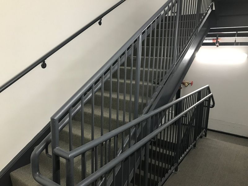 Gray metal staircase with handrails in an indoor stairwell with white walls and a bright ceiling light.