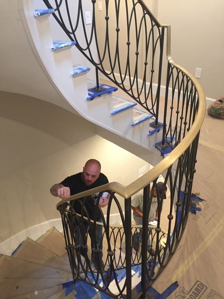 A person installs a wooden handrail on a curved staircase with black metal railings inside a home under construction.