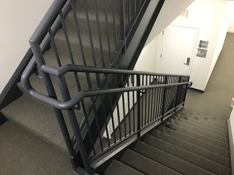 A staircase inside a building with grey carpeted steps and dark grey metal handrails leading to an exit door.