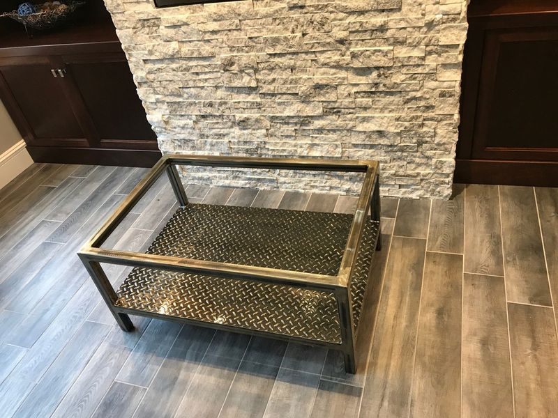 A rectangular metal and glass coffee table with a diamond-plate lower shelf, sitting on a gray wood-look tile floor.