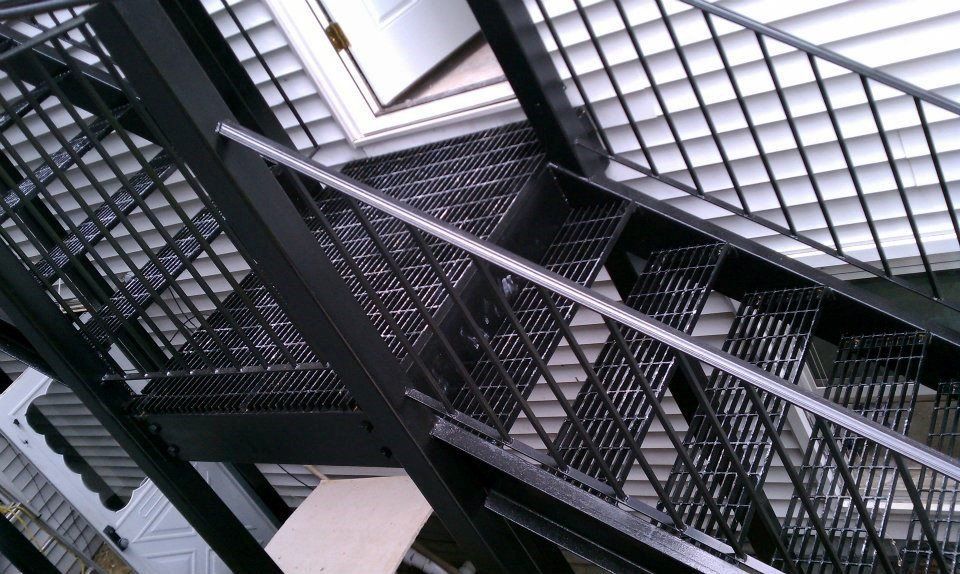 A high-angle view of black metal outdoor stairs and a platform, showing a grate-style walking surface and railings.