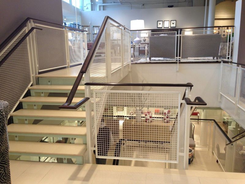 An indoor staircase with white steps and handrails featuring white metal mesh panels, leading to an upper store level.