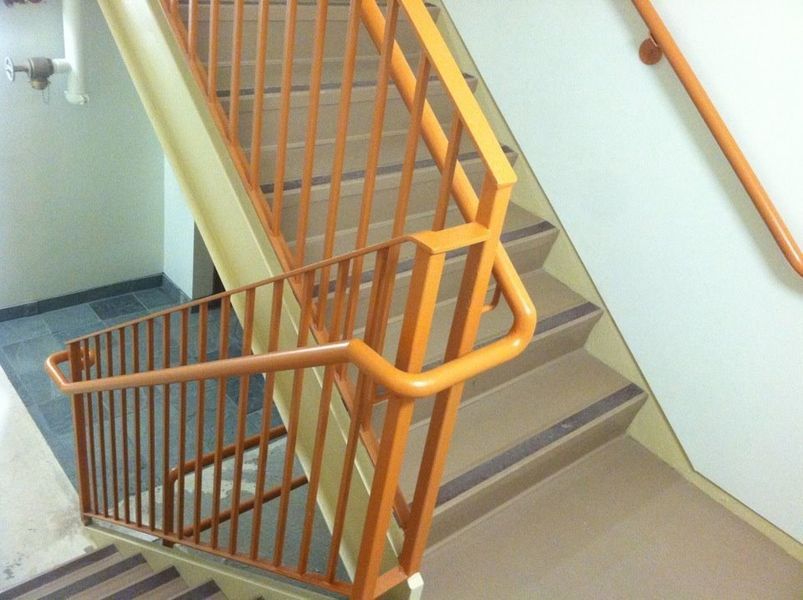 Tan-colored stairs with metal railings, featuring a handrail on the adjacent white wall in an indoor stairwell.