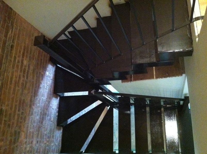 A steep, L-shaped staircase with dark metal steps and railings set against a brick wall.