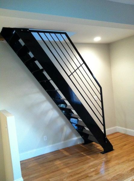 Modern black steel staircase with a matching railing, set against a neutral wall in a room with light wood floors.
