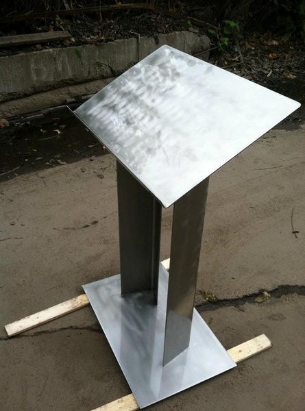 A minimalist metal lectern or podium with a rectangular base and slanted top, standing outdoors on wooden slats.