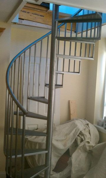 A spiral staircase made of grey metal sits inside a room, leading up to an opening in the ceiling.