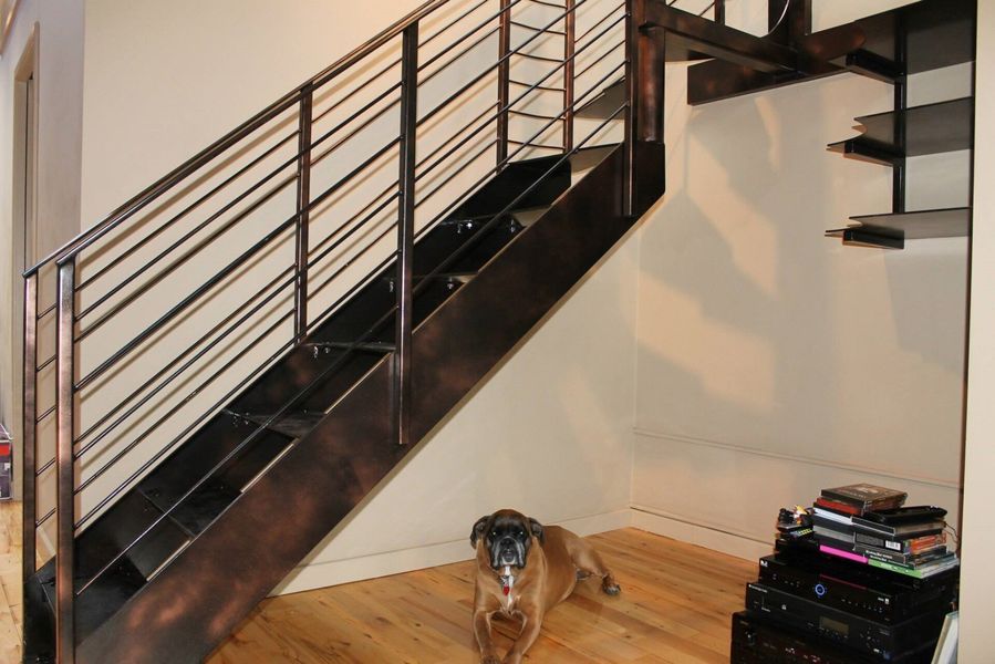 A brown dog lies on a wooden floor in front of a dark metal staircase with horizontal railing and nearby wall shelving.