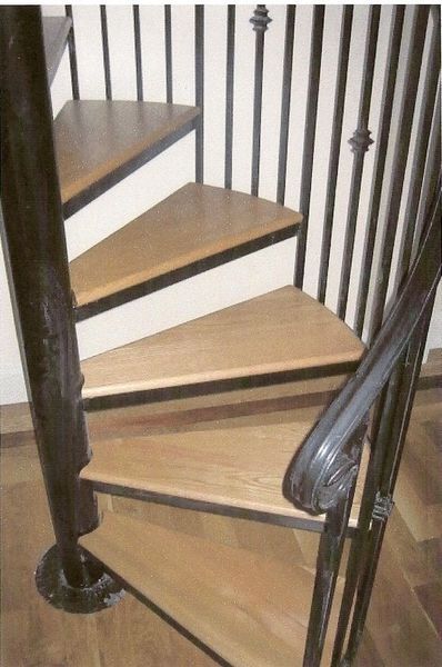 A spiral staircase with light wood steps, dark metal handrails, and a central metal pole, viewed from above.
