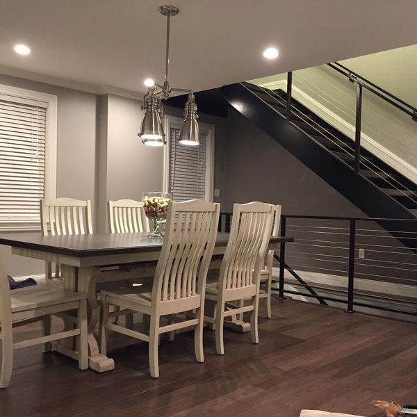 A dining room with a long table, six white spindle-back chairs, and an industrial-style chandelier by a modern staircase.