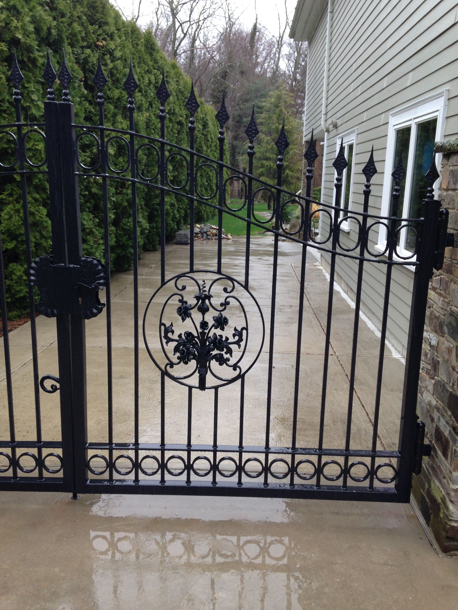 Black wrought-iron gate with floral design beside a concrete driveway and house exterior.