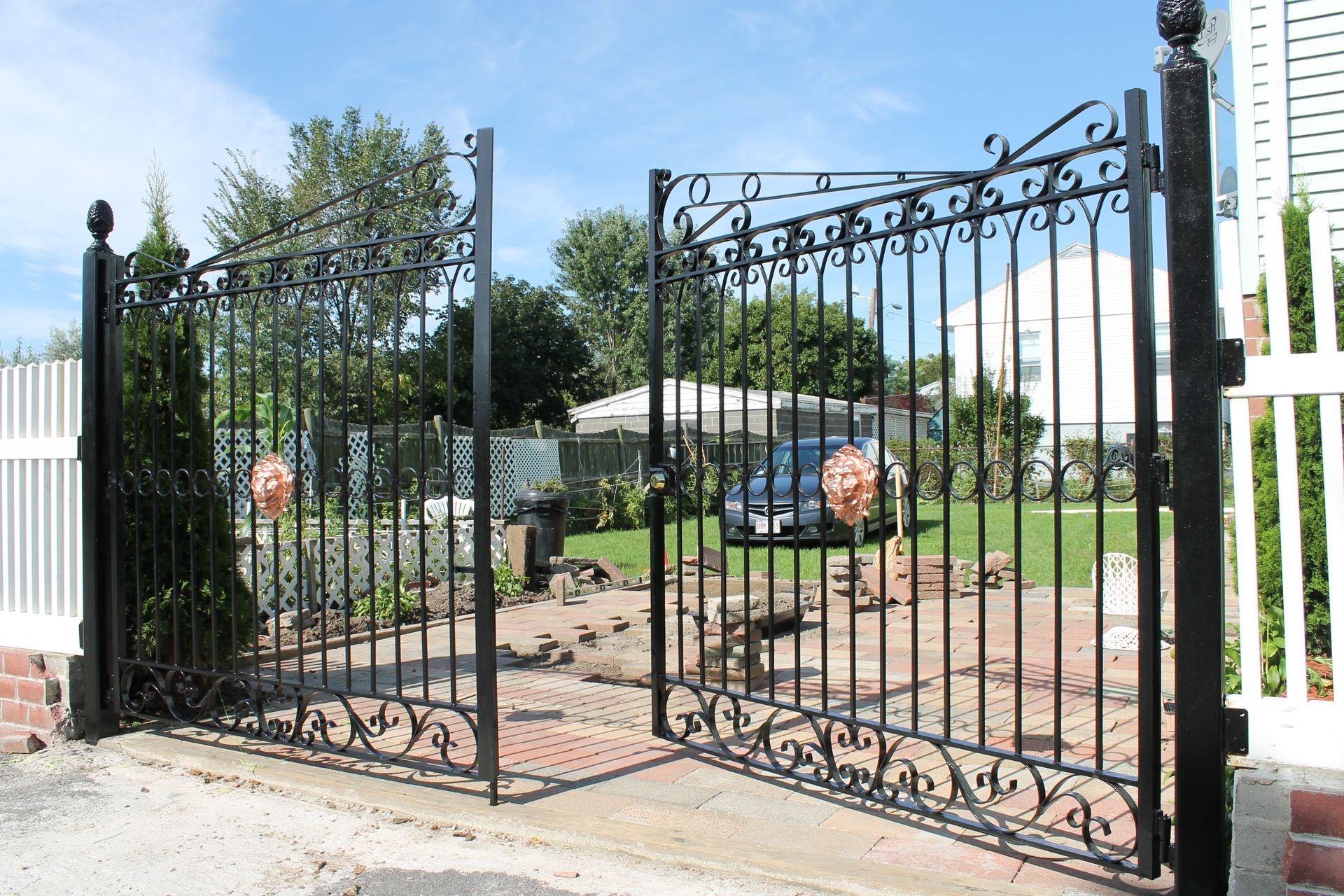 Black wrought-iron driveway gate between white fences under a bright blue sky