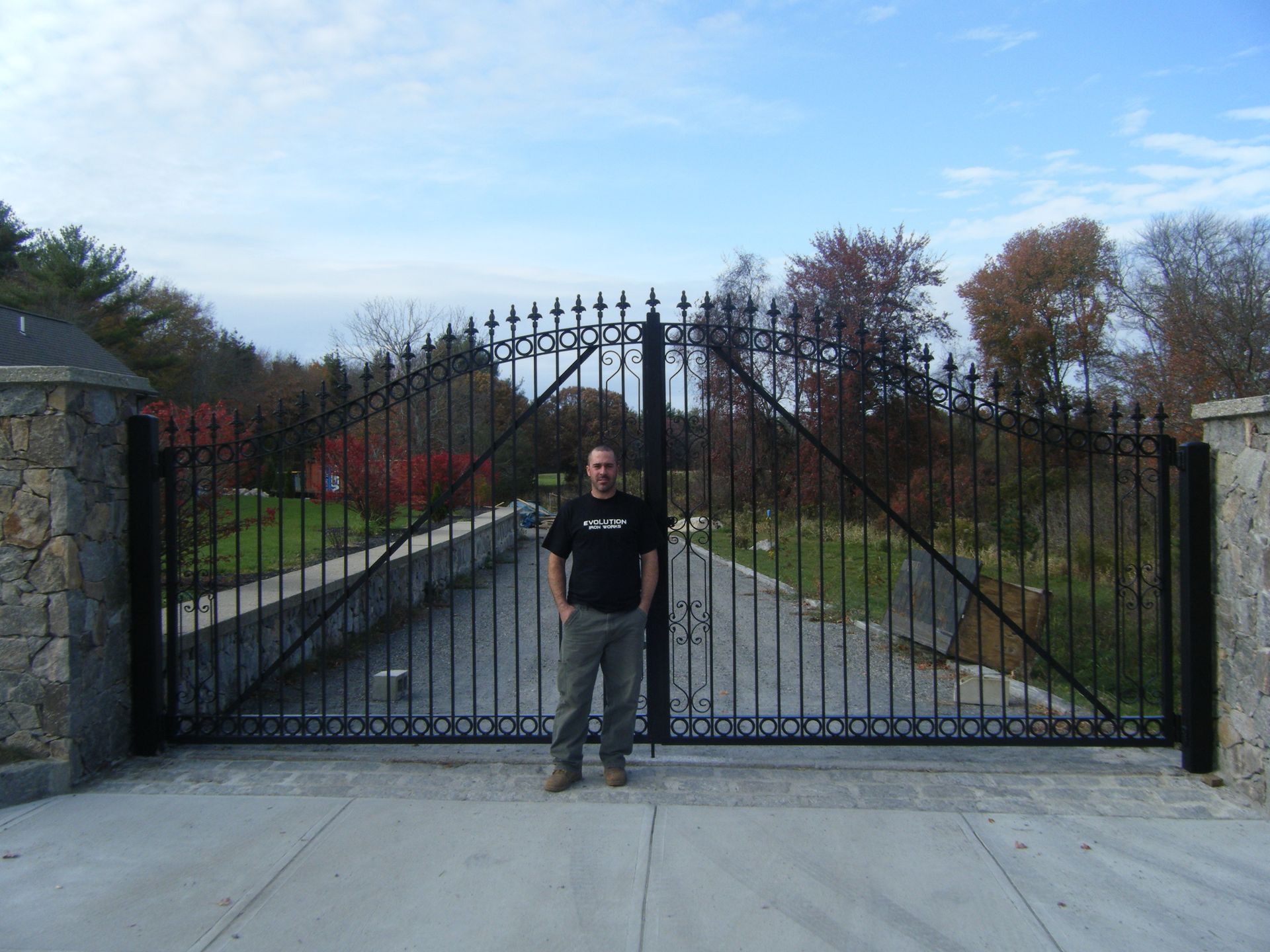 Person standing in front of a large black wrought-iron gate with stone pillars and a driveway behind it.