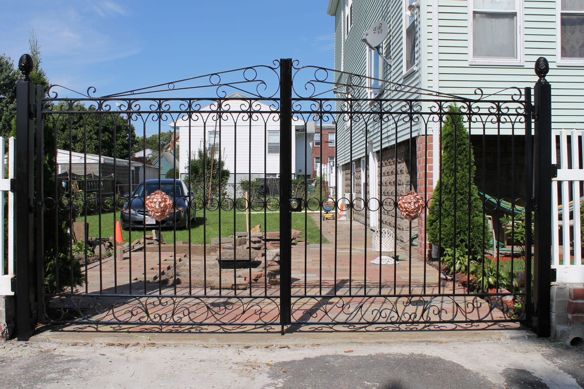 Black wrought-iron gate blocking a narrow front walkway beside a light blue house and white fence.
