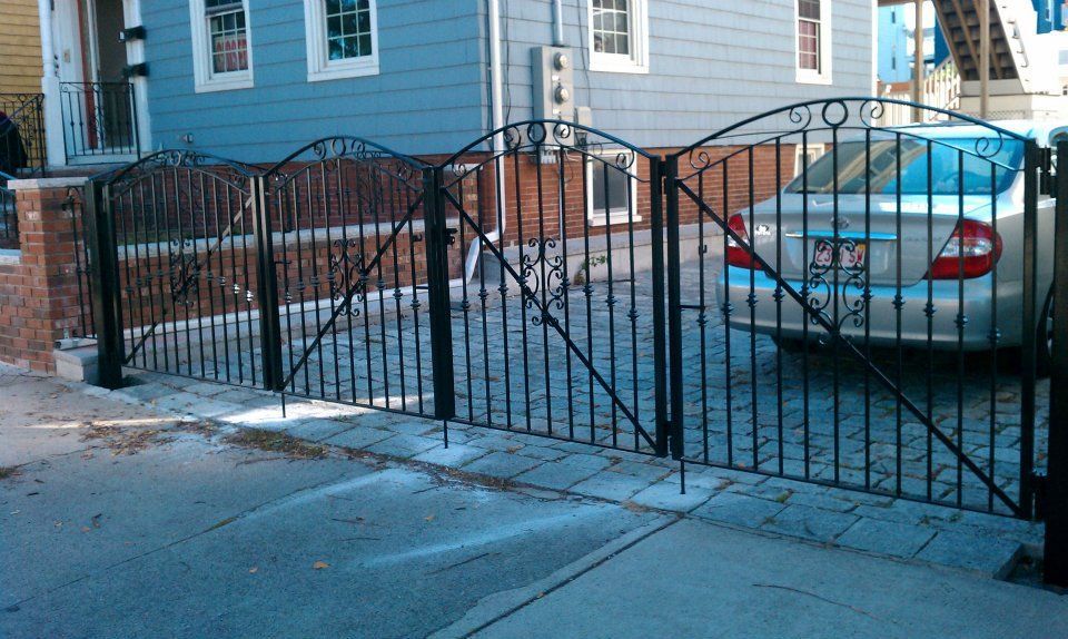 Black metal driveway gate between brick pillars in front of a house, with a parked silver car.