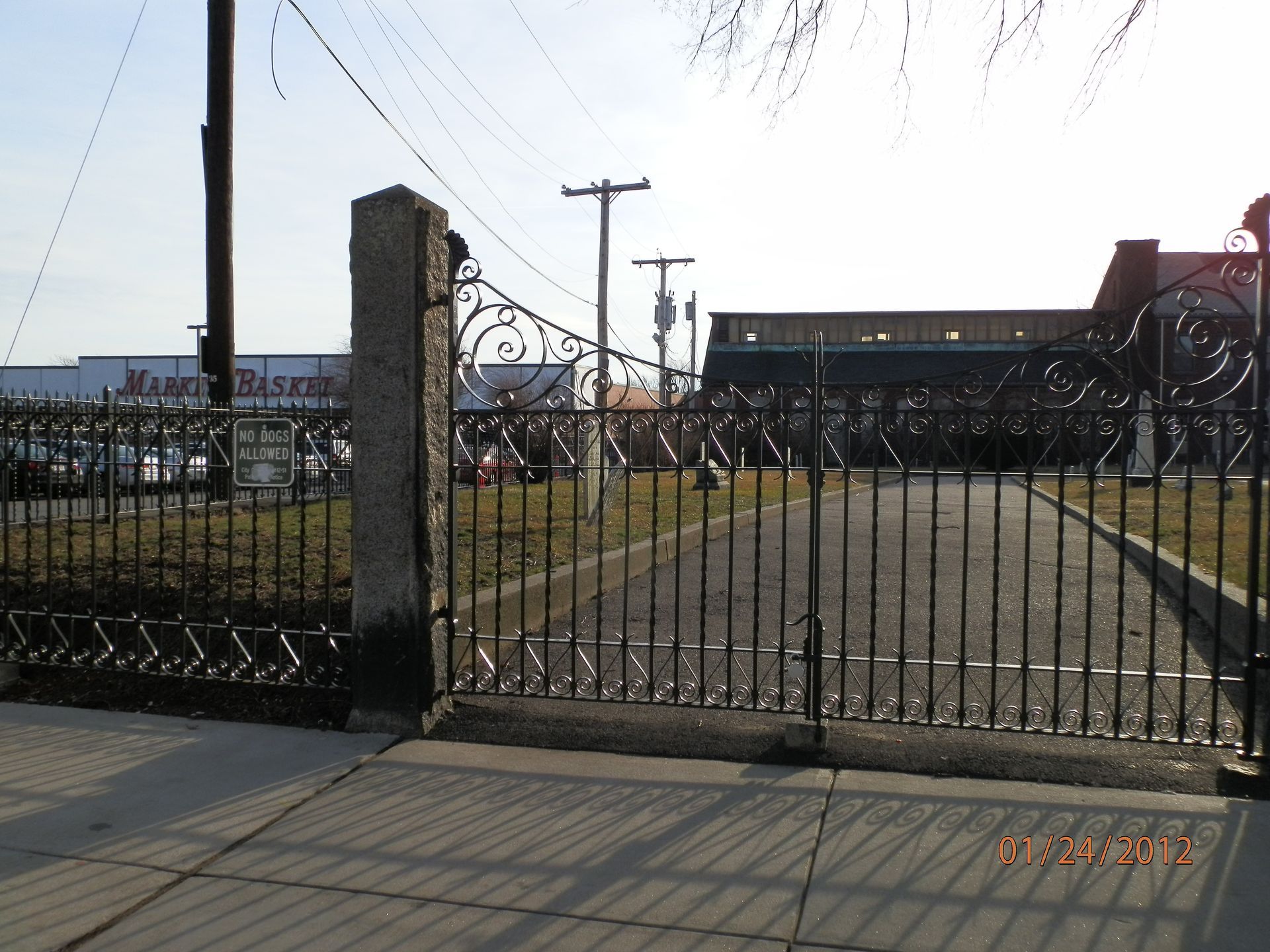 Gated entrance to an industrial site with a paved driveway, metal fence, and buildings in the background