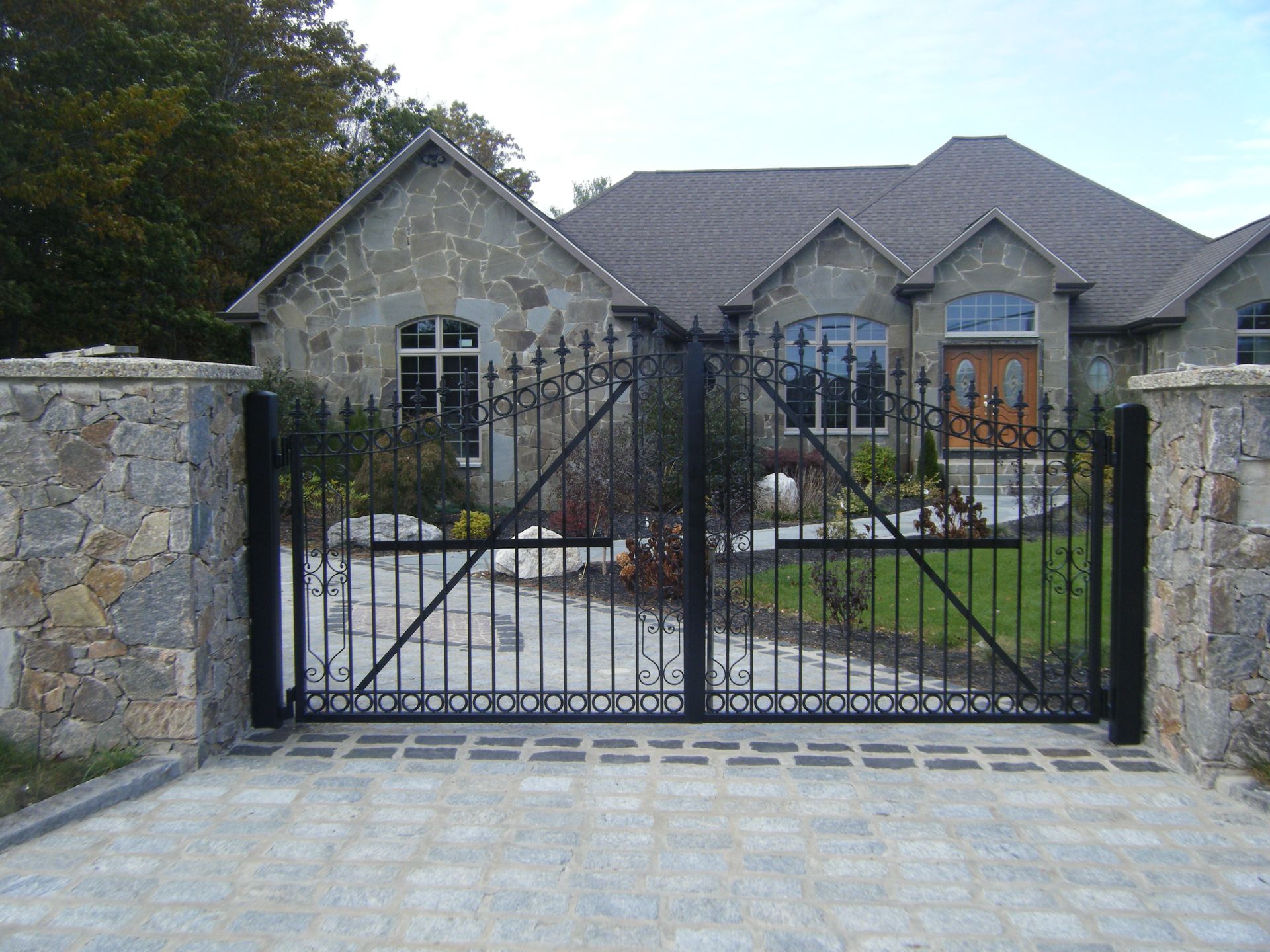 Stone house behind black wrought-iron gate and stone wall at a driveway entrance