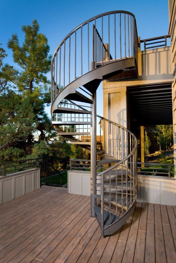 Spiral staircase on a wooden deck beside a house, with trees and blue sky in the background