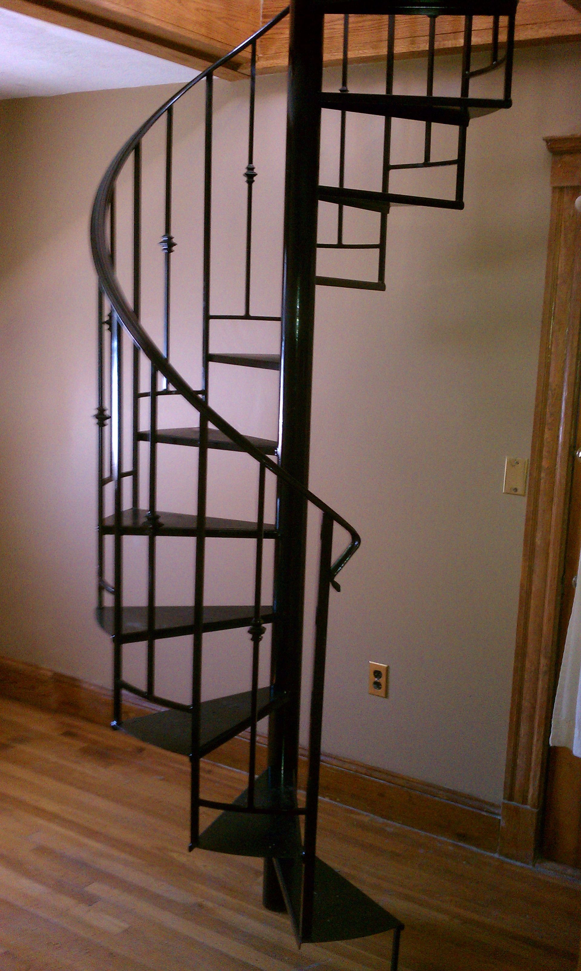 Curved black metal spiral staircase indoors beside a wall and wooden floor