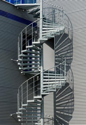 Outdoor metal spiral staircase against a corrugated building wall, with curved railings and shadows.