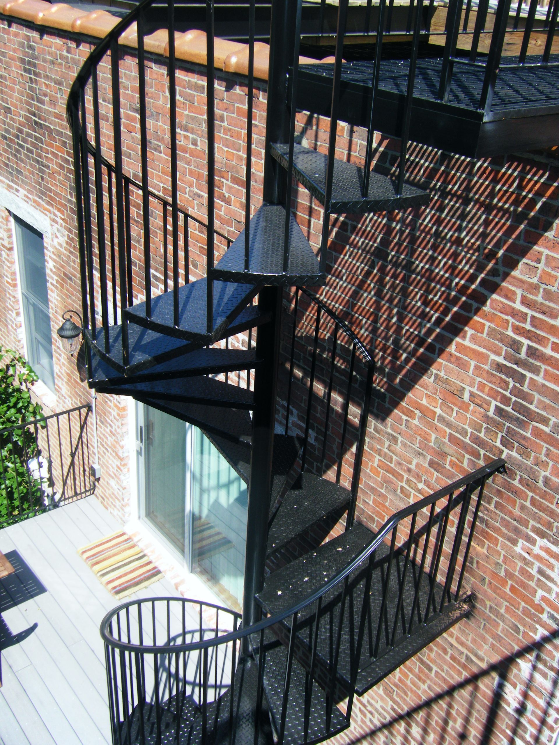 Black metal spiral staircase beside a brick building, casting shadows over a small balcony.