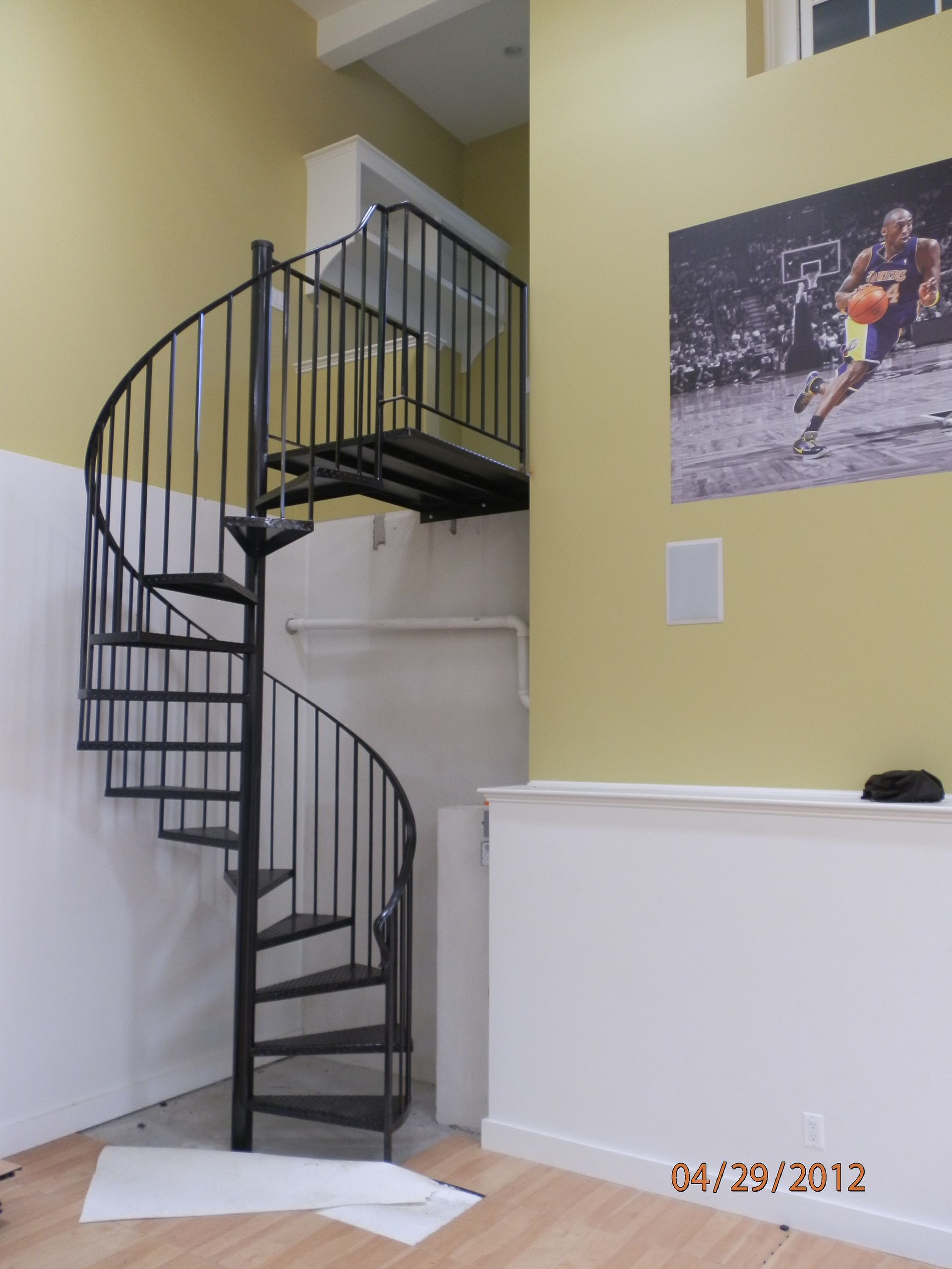 Curved black spiral staircase in a yellow-and-white interior hallway, with a framed sports poster on the wall.