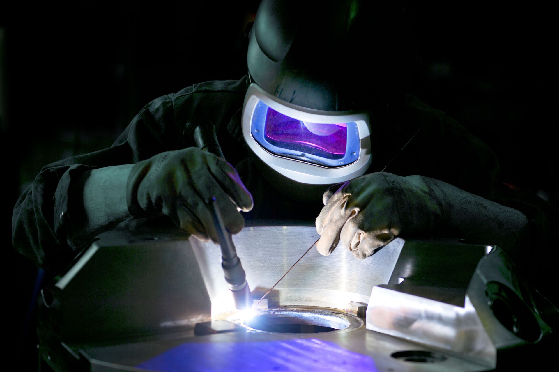 A welder in protective gear uses a TIG torch to join metal, creating a bright blue glow in a dark workshop.