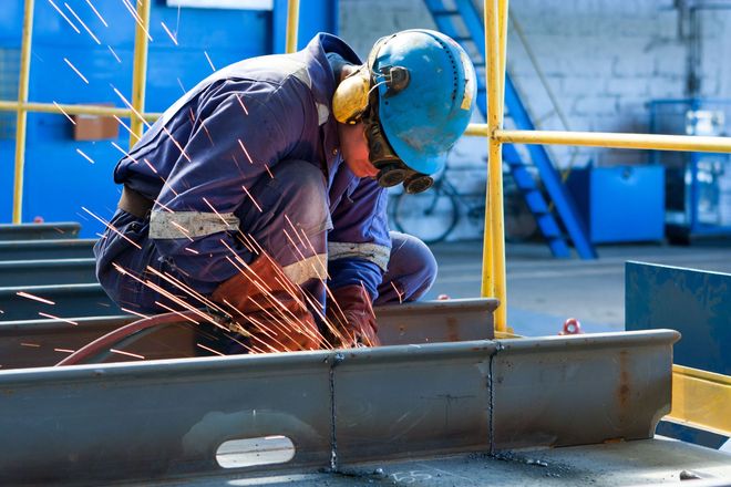 A worker in a blue uniform, helmet, and protective gear uses a grinder to cut a metal beam, creating a spray of sparks.