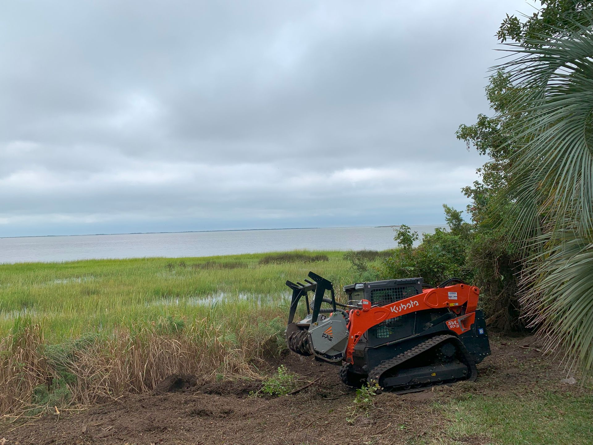 Orange and black track loader clearing brush near water on a cloudy day.