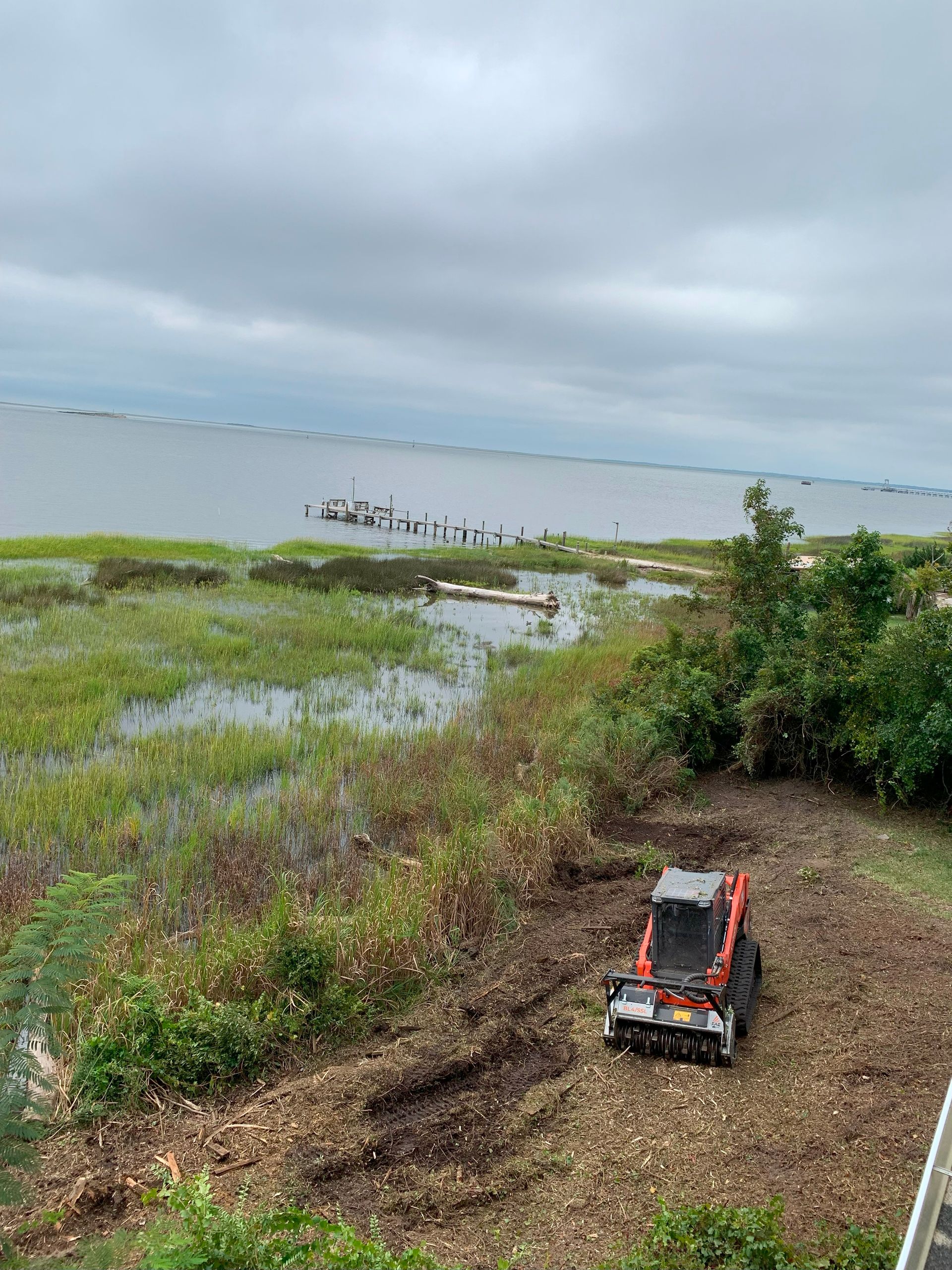 Orange tractor clearing brush near water under a cloudy sky.