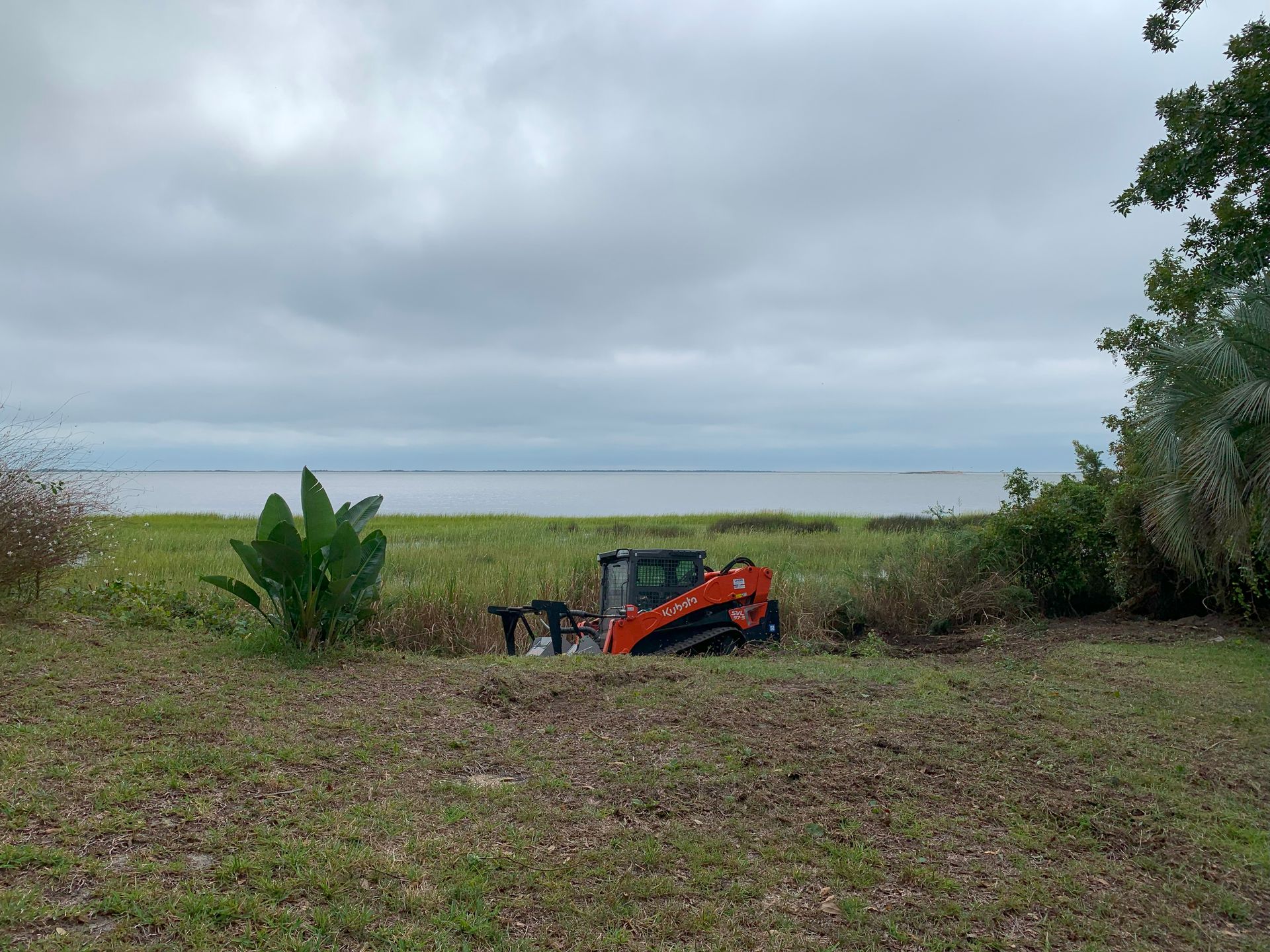 Skid steer clearing brush near water under an overcast sky.