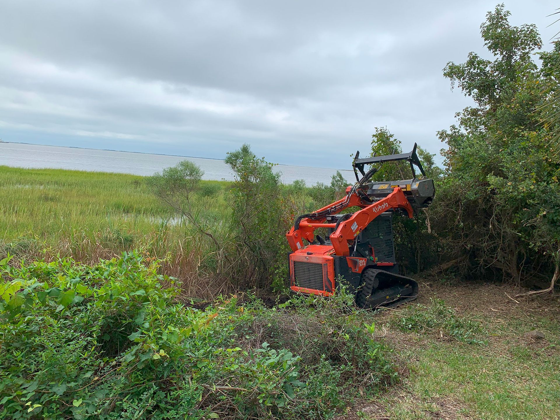 Orange skid steer clearing brush near a body of water under cloudy skies.