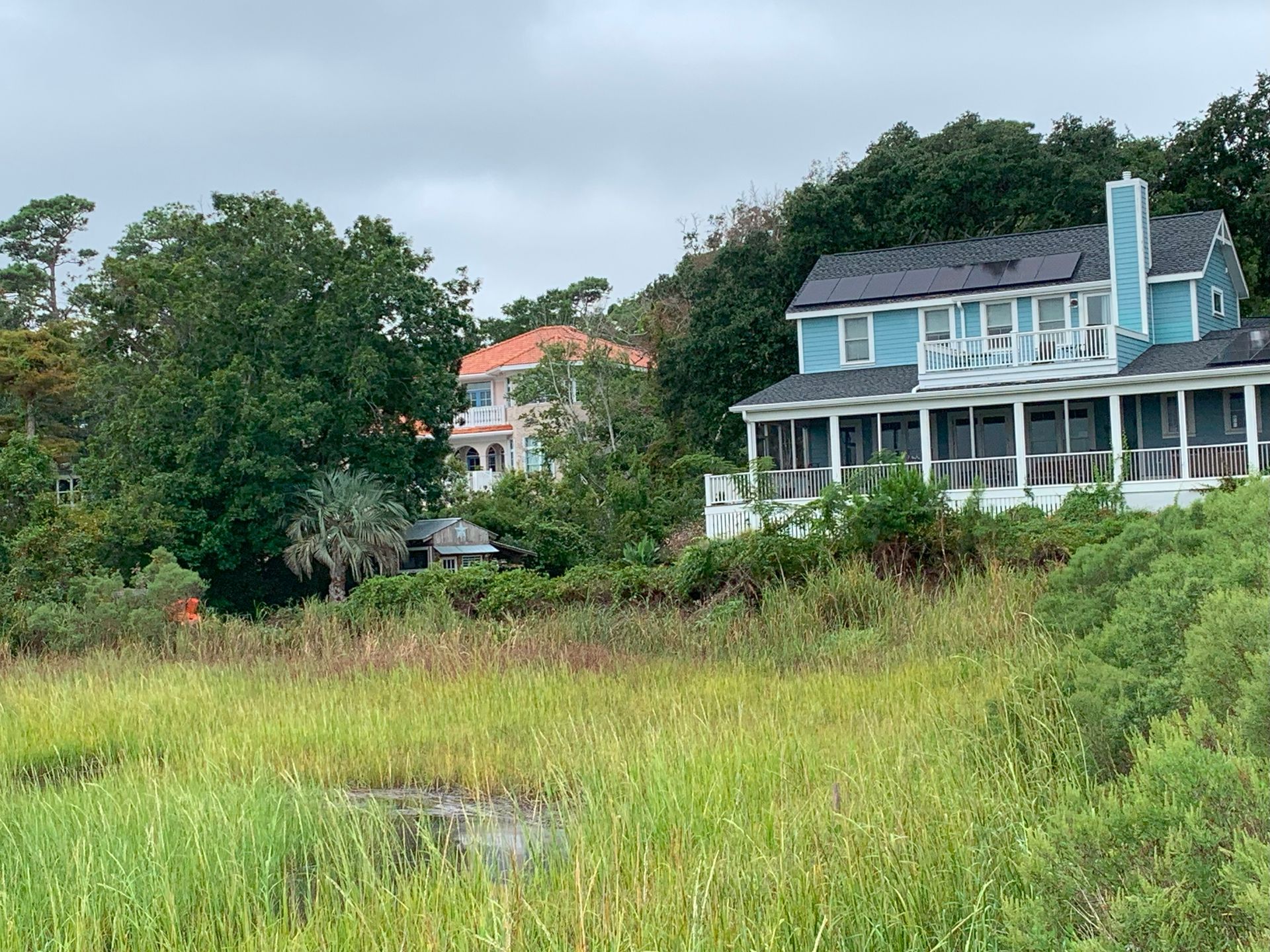 Houses by a marsh with green grass, trees, and a cloudy sky.