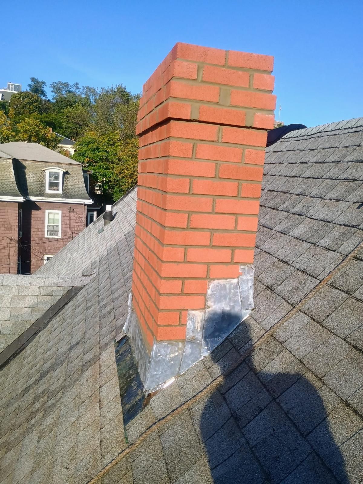 Brick chimney on a shingled roof, set against a blue sky, with the shadow of a person.