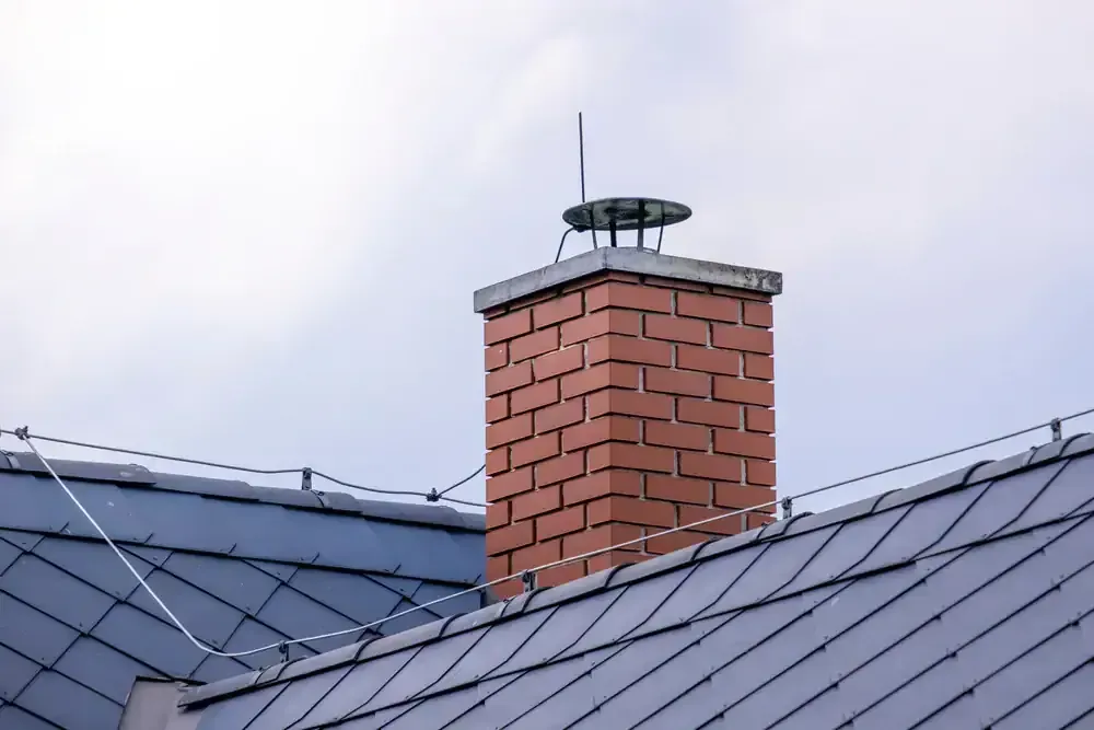 Brick chimney on a gray-roofed building, topped with a metal cap and lightning rod, against a cloudy sky.