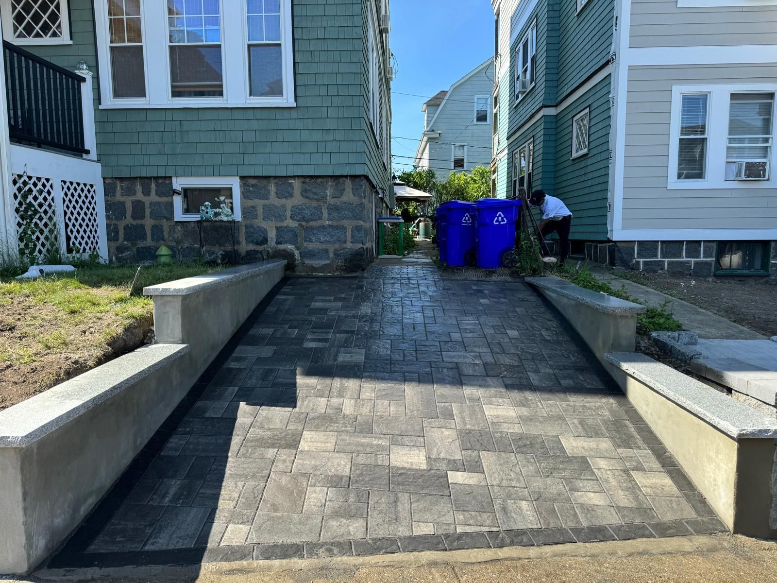 Paver driveway between two buildings, with concrete walls on either side. Blue trash bins visible.