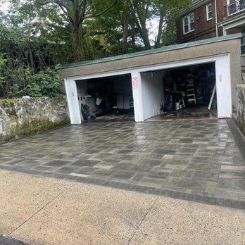 Paver driveway leads to two open garage bays. Garage is attached to a brick building, set on a hillside.