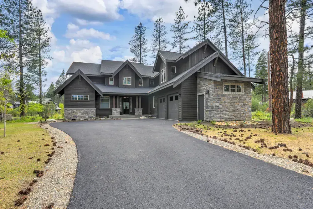 Dark grey house with stone accents, a long driveway, and a forest setting.